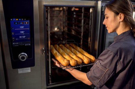 Foto de Mujer panadero usando horno eléctrico para hornear baguettes francesas en panadería - Imagen libre de derechos