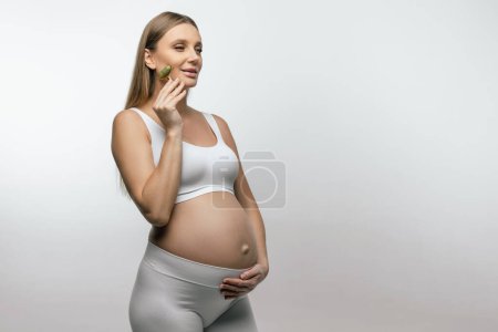 Téléchargez les photos : Procédures beauté. Jolie jeune femme enceinte avec un rouleau de visage dans les mains - en image libre de droit