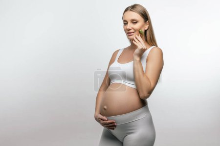 Téléchargez les photos : Procédures beauté. Jolie jeune femme enceinte avec un rouleau de visage dans les mains - en image libre de droit
