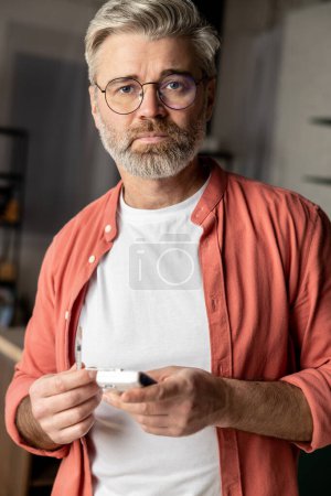 Téléchargez les photos : Homme diabétique aux cheveux gris tenant vérifier la glycémie glucomètre à l'intérieur de la maison - en image libre de droit
