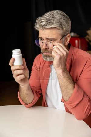 Foto de Barbudo hombre de mediana edad leyendo detrás de un frasco de medicina en casa - Imagen libre de derechos