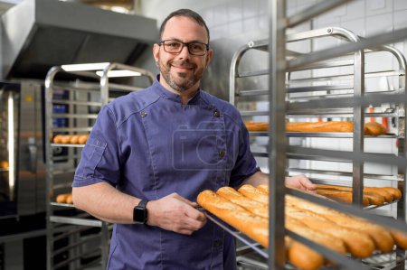 Téléchargez les photos : Boulanger professionnel près de pain frais cuit sur chariot porte-plateau dans l'atelier de boulangerie - en image libre de droit