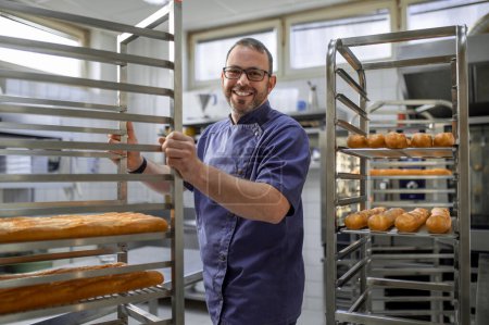 Photo for Professional man baker near fresh baked bread on tray rack trolley in bakery workshop - Royalty Free Image