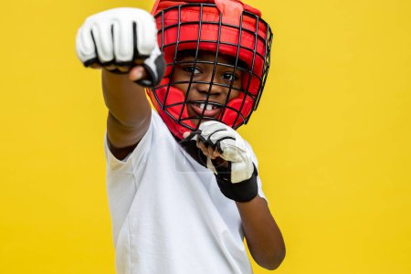 Téléchargez les photos : Petit garçon combattant à la peau foncée portant un casque de boxe protecteur et des gants isolés sur fond jaune - en image libre de droit