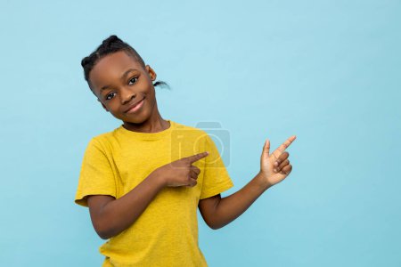 Téléchargez les photos : Petit garçon afro-américain souriant pointant vers l'espace de copie pour la publicité isolée sur fond bleu - en image libre de droit