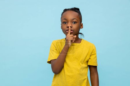Téléchargez les photos : Sérieux petit garçon à la peau foncée montrant un geste de silence isolé sur fond bleu - en image libre de droit