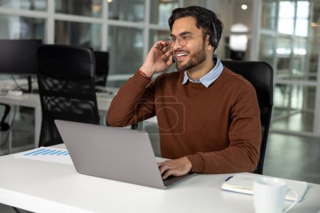 Téléchargez les photos : Homme barbu adulte employé de bureau avec casque de travail sur ordinateur portable dans un bureau moderne - en image libre de droit