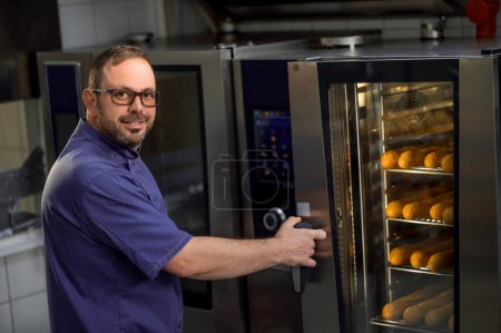 Téléchargez les photos : Boulanger professionnel dans la cuisine près du four avec baguettes dans la boulangerie - en image libre de droit