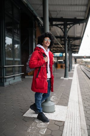 Téléchargez les photos : J'attends le train. Guy en veste rouge et avec sac à dos attendant le train au quai - en image libre de droit