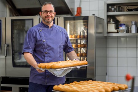 Téléchargez les photos : Homme boulanger en uniforme baguettes de cuisson avec étagères à pain sur le fond à la fabrication - en image libre de droit