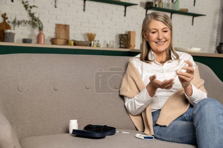 Téléchargez les photos : Test de glycémie. Belle femme d'âge moyen assise sur le canapé et faisant un test de glycémie - en image libre de droit
