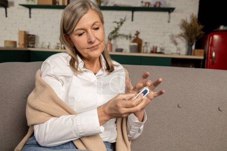 Photo for Diabetic woman. Mature woman sitting on the sofa and checking blood sugar with glucometer - Royalty Free Image