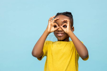 Téléchargez les photos : Drôle sombre peau petit garçon regardant avec mains jumelles isolé sur fond bleu - en image libre de droit