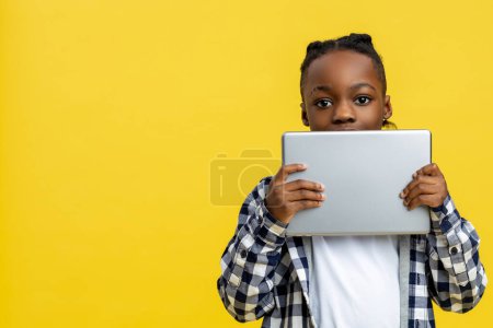 Foto de Un chico con un aparato. Niño afroamericano en camisa a cuadros con tableta en las manos - Imagen libre de derechos