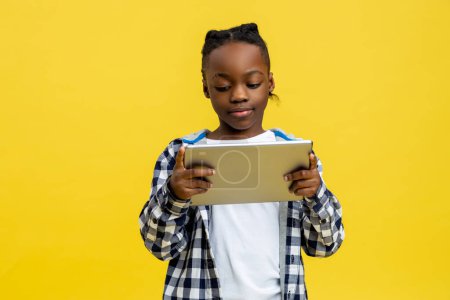 Foto de Un chico con un aparato. Niño afroamericano en camisa a cuadros con tableta en las manos - Imagen libre de derechos