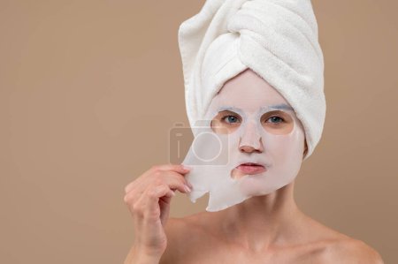 Attraktive Frau mit Kosmetikmaske im Gesicht, die isoliert vor braunem Hintergrund steht