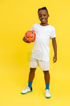 Photo for Full length of Afro-American little boy wearing white T-shirt posing with ball isolated over yellow background - Royalty Free Image