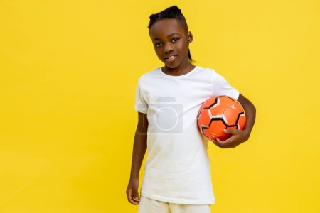 Foto de Atlético chico afroamericano en uniforme con pelota de fútbol aislado sobre fondo amarillo - Imagen libre de derechos