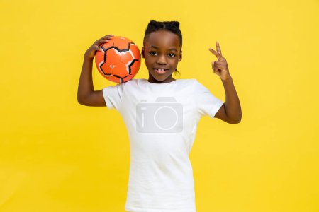 Foto de Pequeño niño alegre afroamericano jugando fútbol en el campeonato de niños mostrando v signo aislado sobre fondo amarillo - Imagen libre de derechos