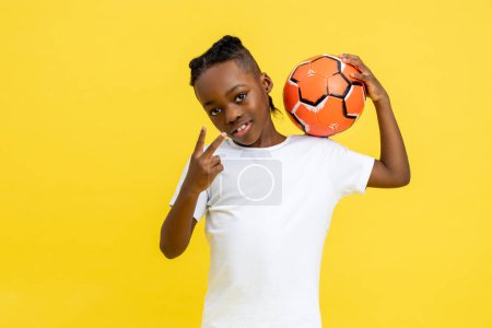 Foto de Niño afroamericano con camiseta blanca posando con pelota mostrando signo de victoria aislado sobre fondo amarillo - Imagen libre de derechos