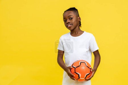 Photo for Afro-American little boy wearing white T-shirt posing with ball isolated over yellow background, copy space - Royalty Free Image