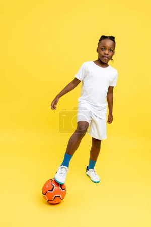 Foto de Longitud completa de lindo niño afroamericano de pie con pelota de fútbol en el entrenamiento aislado sobre fondo amarillo - Imagen libre de derechos