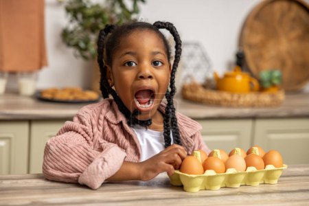 Téléchargez les photos : Petite fille noire mignonne avec expression faciale excitée assise dans la cuisine avec des œufs sur le comptoir - en image libre de droit