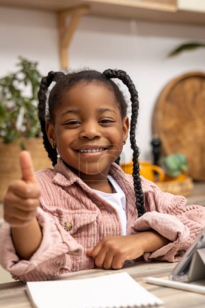 Photo for Happy black schoolgirl learning at home with textbook sitting at desk looking at camera doing lessons showing like gesture - Royalty Free Image