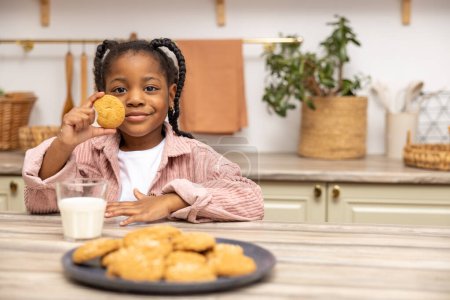 Photo for Cute little black girl sitting at table having snack milk and cookies in kitchen interior - Royalty Free Image