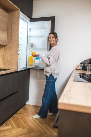 In the kitchen. Smiling contented woman standing near the fridge in the kitchen