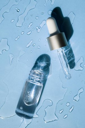 Foto de Botella de vidrio y pipeta en un líquido transparente cosmético sobre un fondo azul. Puesta plana. - Imagen libre de derechos