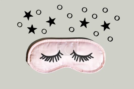 Pinkfarbene Schlafmaske mit Wimpern auf grauem Hintergrund mit schwarzen Sternen. Gesunder Schlaf. Platz für Text, Draufsicht.