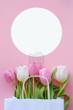 Photo for Pink tulips in a white paper bag and a white circle for an inscription or congratulations on a pink background. Vertical image, flat lay, copy space. - Royalty Free Image