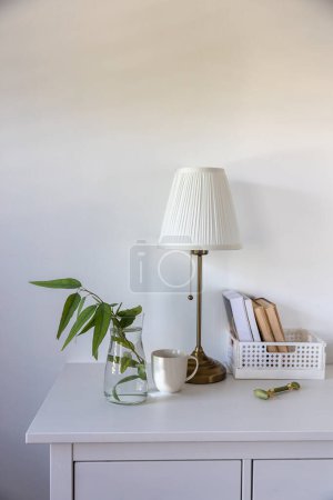 Photo for A branch of eucalyptus in a glass vase, a box of books, a massage roller for the face, a cup of coffee on a white chest of drawers against the wall. Ready layout. Vertical frame. Space for text - Royalty Free Image