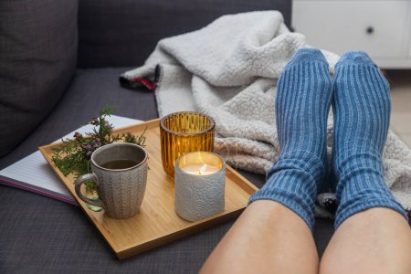 Photo for Soft photo of a woman`s legs in woolen blue socks on the sofa with a book and cup of tea and a candle on the tray. Interior and home coziness concept. Top view - Royalty Free Image