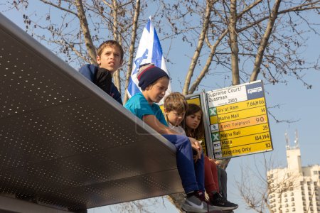 Foto de JERUSALEM, ISRAEL - February 20 2023: Israelis protest near the Knesset against plans by prime minister Benjamin Netanyahu. Children with flags sit on the roof of the bus stop - Imagen libre de derechos
