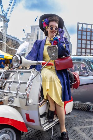 Foto de Londres Reino Unido - 15 de abril de 2023: Classic Car Boot Sale by Vintage. Festival retro. Personas que venden ropa vintage, joyas, artículos para el hogar en sus vehículos clásicos. Chica en una scooter Vespa. - Imagen libre de derechos