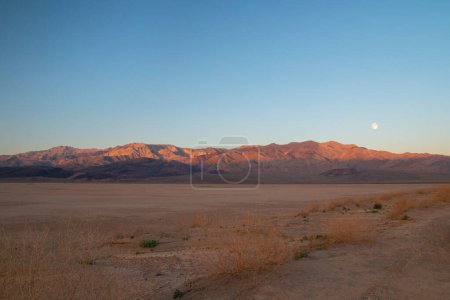Foto de Amanecer luz de la mañana en el prístino paisaje desierto de sal plana con montañas de color rojo dorado y la luna llena por encima, Panamint Valley en el Parque Nacional del Valle de la Muerte California - Imagen libre de derechos