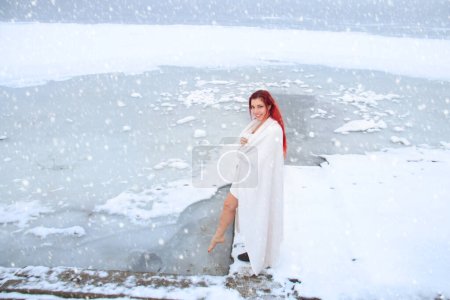 Foto de Mujer envuelta en una toalla probando agua fría con los pies en el paisaje helado del lago mientras cae nieve - Imagen libre de derechos