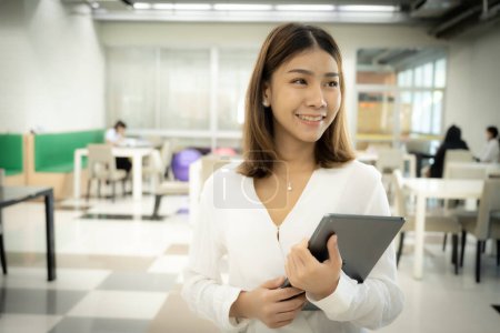 Beautiful asian female teacher is standing while holding tablet with a smiling face in the library room.