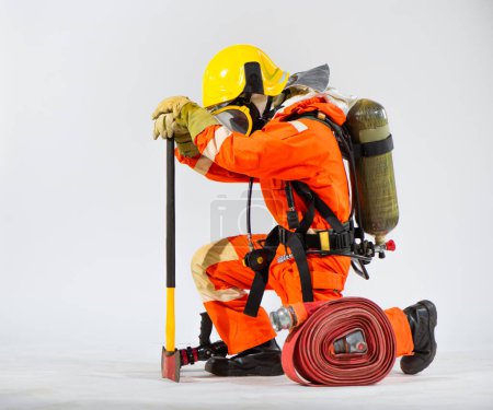Kneeling firefighter presses his axe to the ground in preparation with an oxygen tank on his back against a white background.