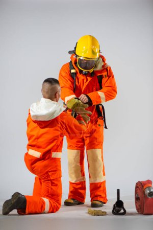 Téléchargez les photos : Un pompier professionnel s'agenouille pour aider son partenaire à enfiler des gants afin d'assurer la sécurité et la préparation à la tâche.. - en image libre de droit
