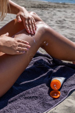 Téléchargez les photos : Une jeune femme applique de la crème solaire sur ses jambes sur une plage de sable, prenant soin de sa peau. Vacances d'été. Gros plan. - en image libre de droit