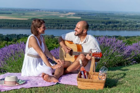 Photo for A young couple in love enjoys a picnic in a beautiful picturesque lavender field by the river. The husband is playing guitar to his lovely wife. - Royalty Free Image