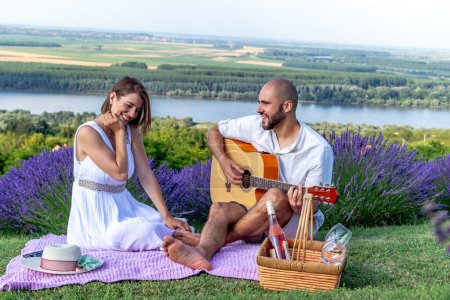 Photo for A young couple in love enjoys a picnic in a beautiful picturesque lavender field by the river. The husband is playing guitar to his lovely wife. - Royalty Free Image