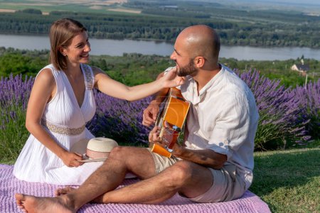 Photo for A romantic couple in love enjoys a picnic in a beautiful picturesque lavender field by the river. The husband is playing guitar to his lovely wife. - Royalty Free Image
