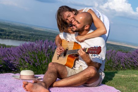 Photo for A romantic couple in love enjoying in a beautiful picturesque lavender field by the river. The husband is playing guitar to his lovely wife. - Royalty Free Image