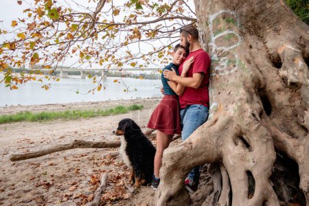 Téléchargez les photos : Un couple amoureux s'étreignant et s'embrassant sous un arbre aux grandes racines croisées et proéminentes, tandis que leur chien de montagne bernois veille. - en image libre de droit