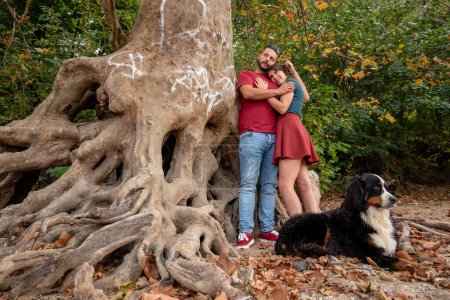 Photo for A couple in love hugging and kissing under a tree with large crisscrossed and prominent roots, while their  Bernese mountain dog is keeping a watch. - Royalty Free Image