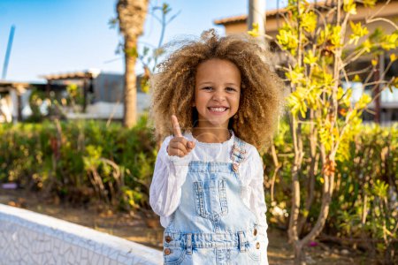 Photo for Close up portrait of a smiling child girl in spring park on sunny warm day pointing finger up. - Royalty Free Image
